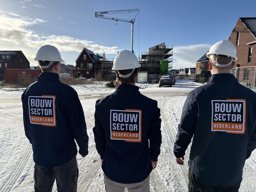 Drie vakmensen in donkerblauwe jassen met het oranje logo van Bouwsector Nederland op de rug bij een bouwplaats
